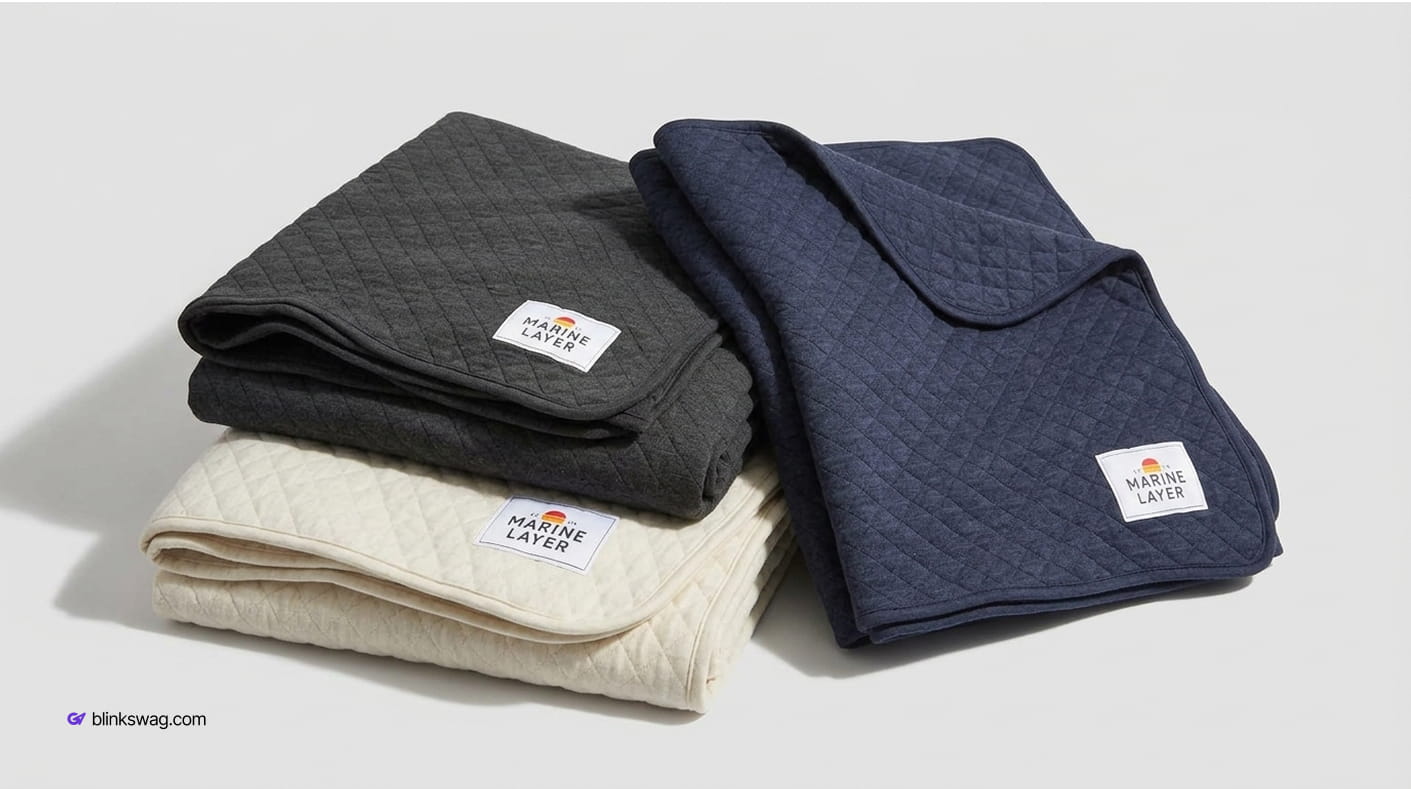 Trio of folded quilted Marine Layer Corbet blankets stacked in charcoal grey, off-white cream, and navy blue.